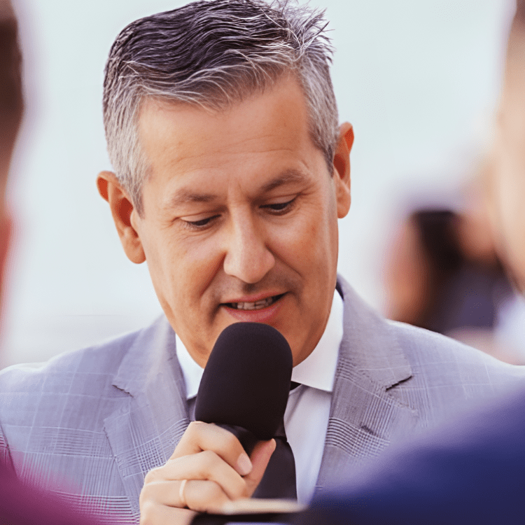 Hombre con traje gris hablando por micrófono en un evento.