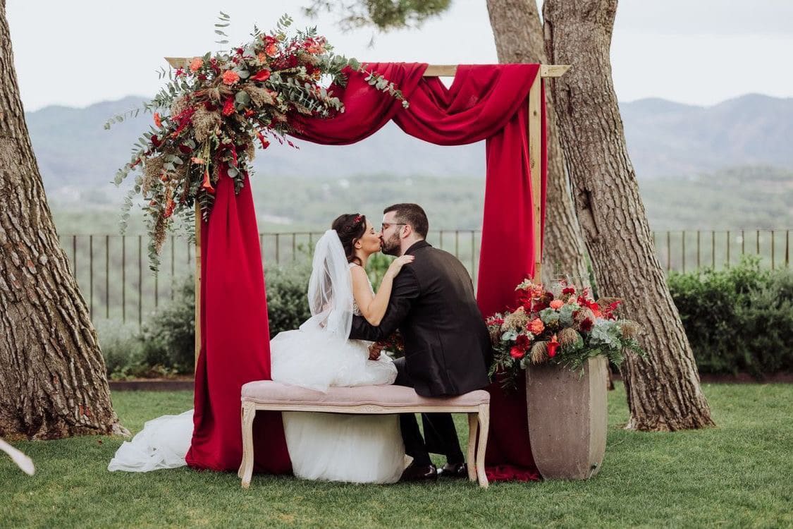 Pareja de novios sentados frente a un altar decorado con tela roja y flores en un jardín al aire libre.