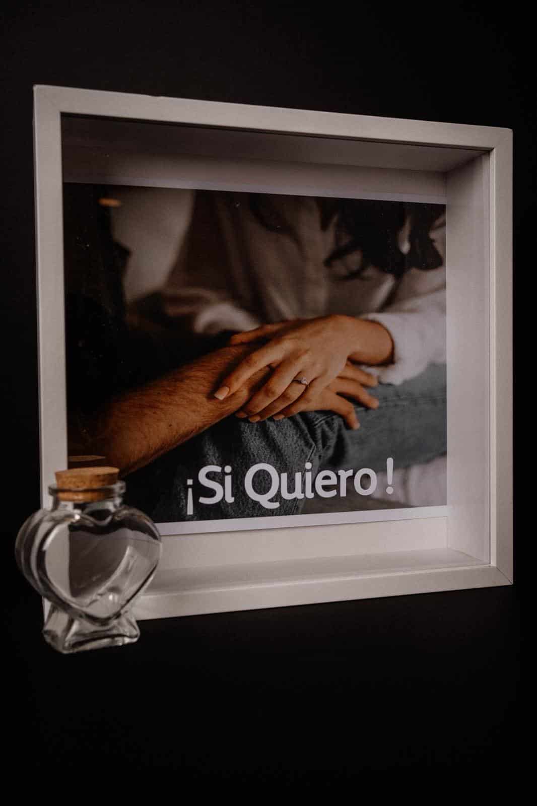 Manos entrelazadas con anillo de compromiso, fondo con texto "¡Sí Quiero!" y frasco decorativo.