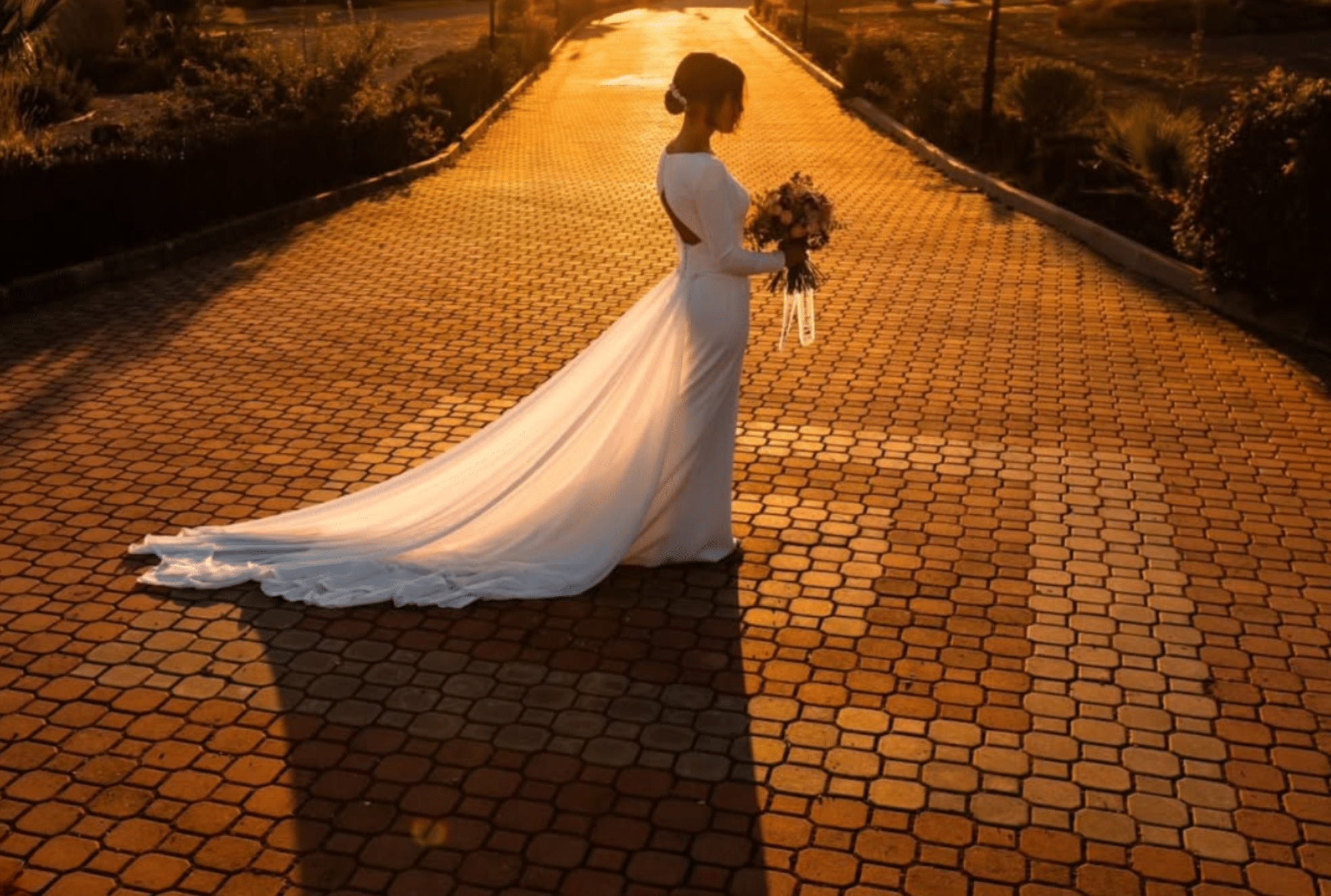 Novia con vestido blanco y ramo, de pie en un camino empedrado al atardecer.