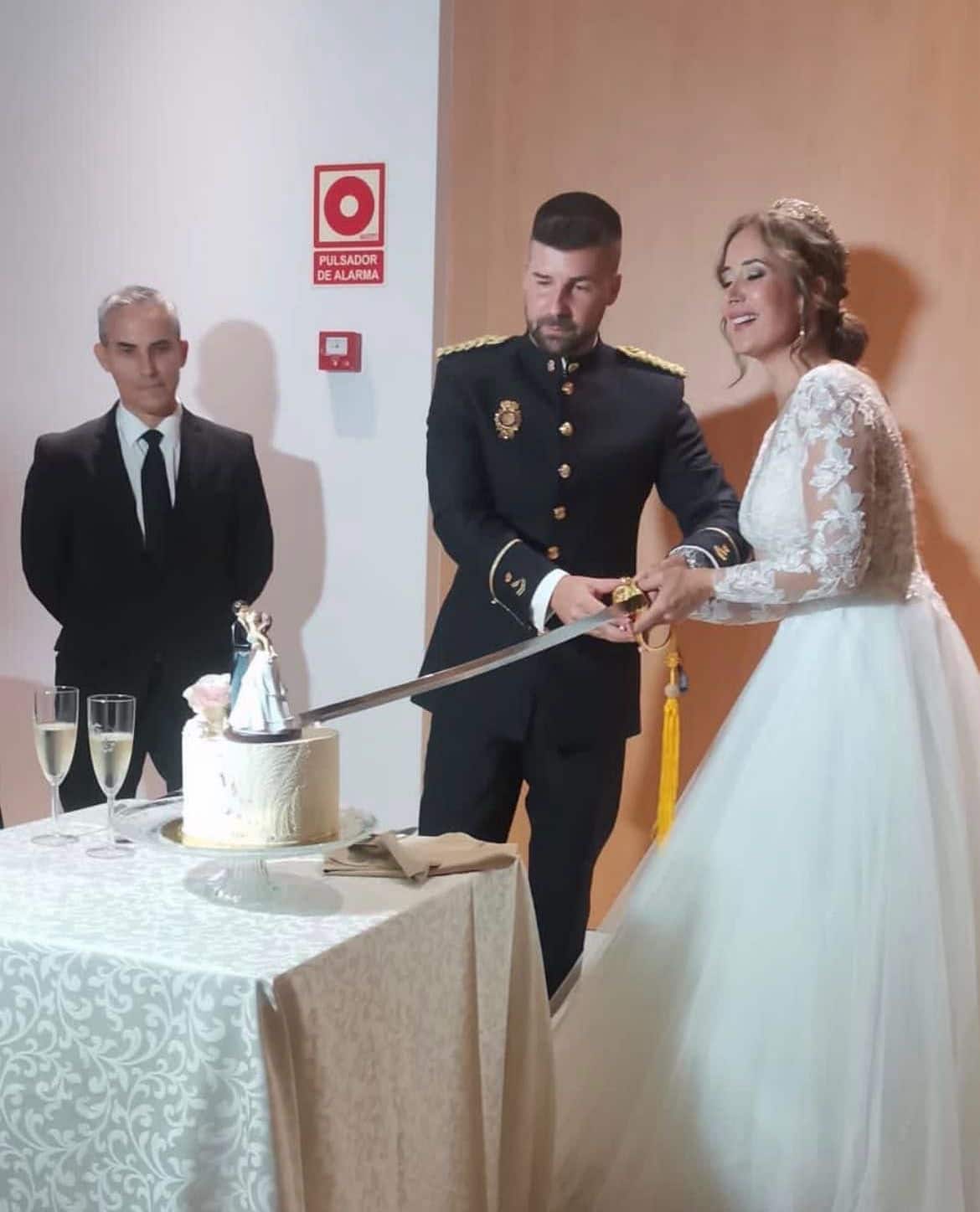Novia y novio cortando la tarta nupcial, junto a un invitado, en un ambiente festivo.