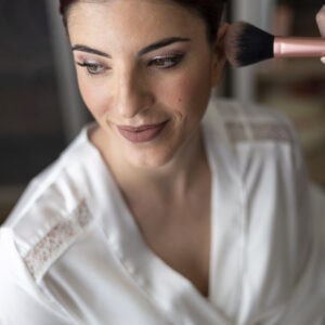 Mujer recibiendo maquillaje, sonriendo mientras un profesional aplica rubor en su rostro.