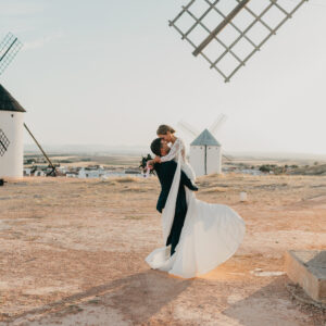 Pareja de novios en un paisaje con molinos, el novio levanta a la novia mientras ella lleva un vestido blanco.