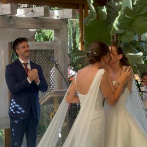 Pareja de novias compartiendo un beso durante la ceremonia, con un oficiante aplaudiendo.