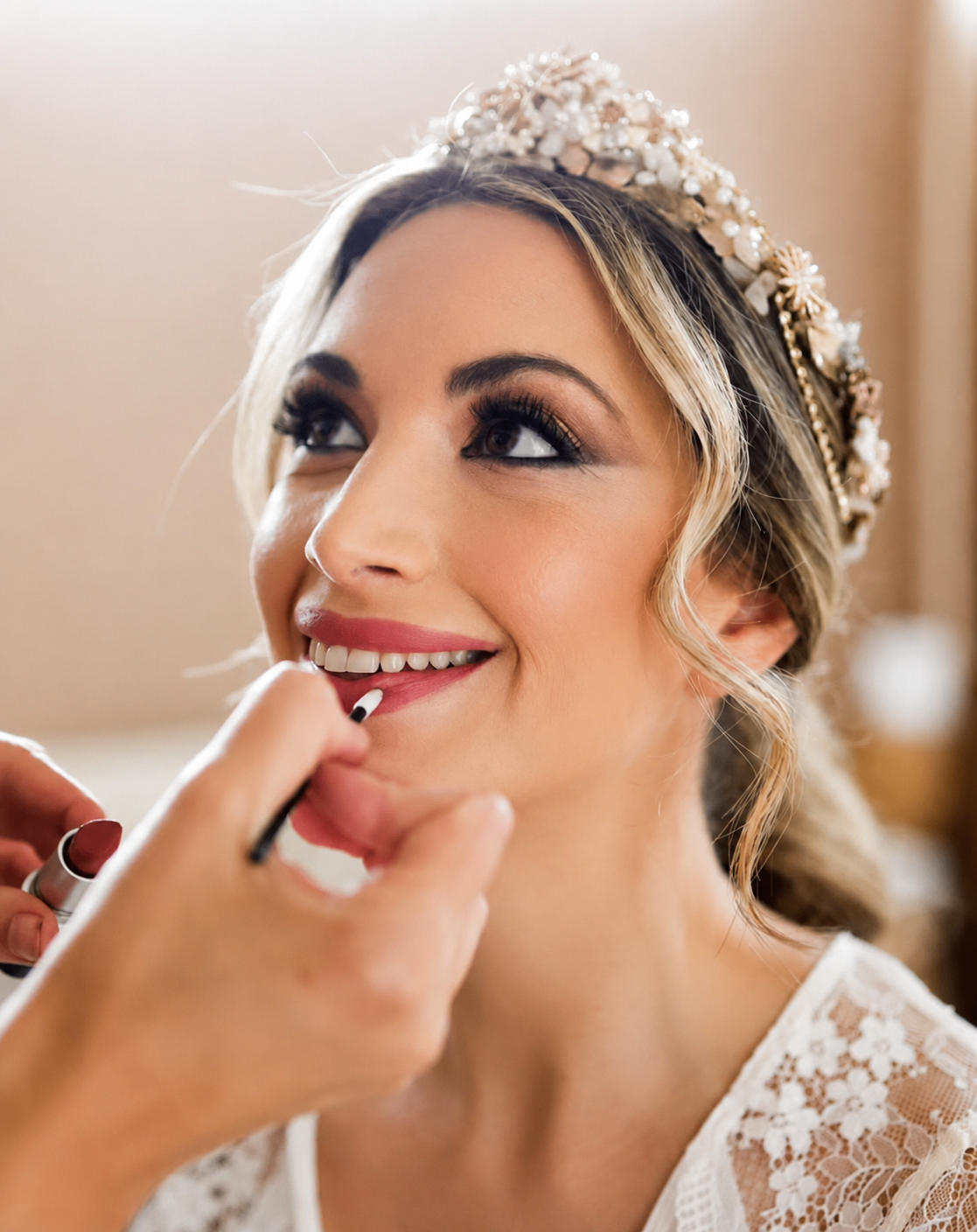 1e421534-0ff0-411d-8d67-5455a174a875 Novia sonriente con maquillaje profesional aplicándose el labial y tocado de flores en el cabello.