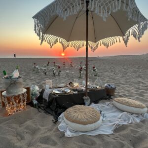 Atardecer en la playa con manta, cojines, mesa baja con comida y sombrilla blanca de macramé.