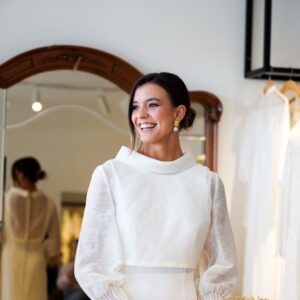 Novia sonriente con vestido blanco y maquillaje natural frente a espejo en habitación luminosa.