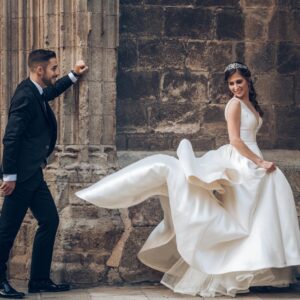 Novios sonrientes en traje y vestido de boda junto a una pared de piedra antigua.