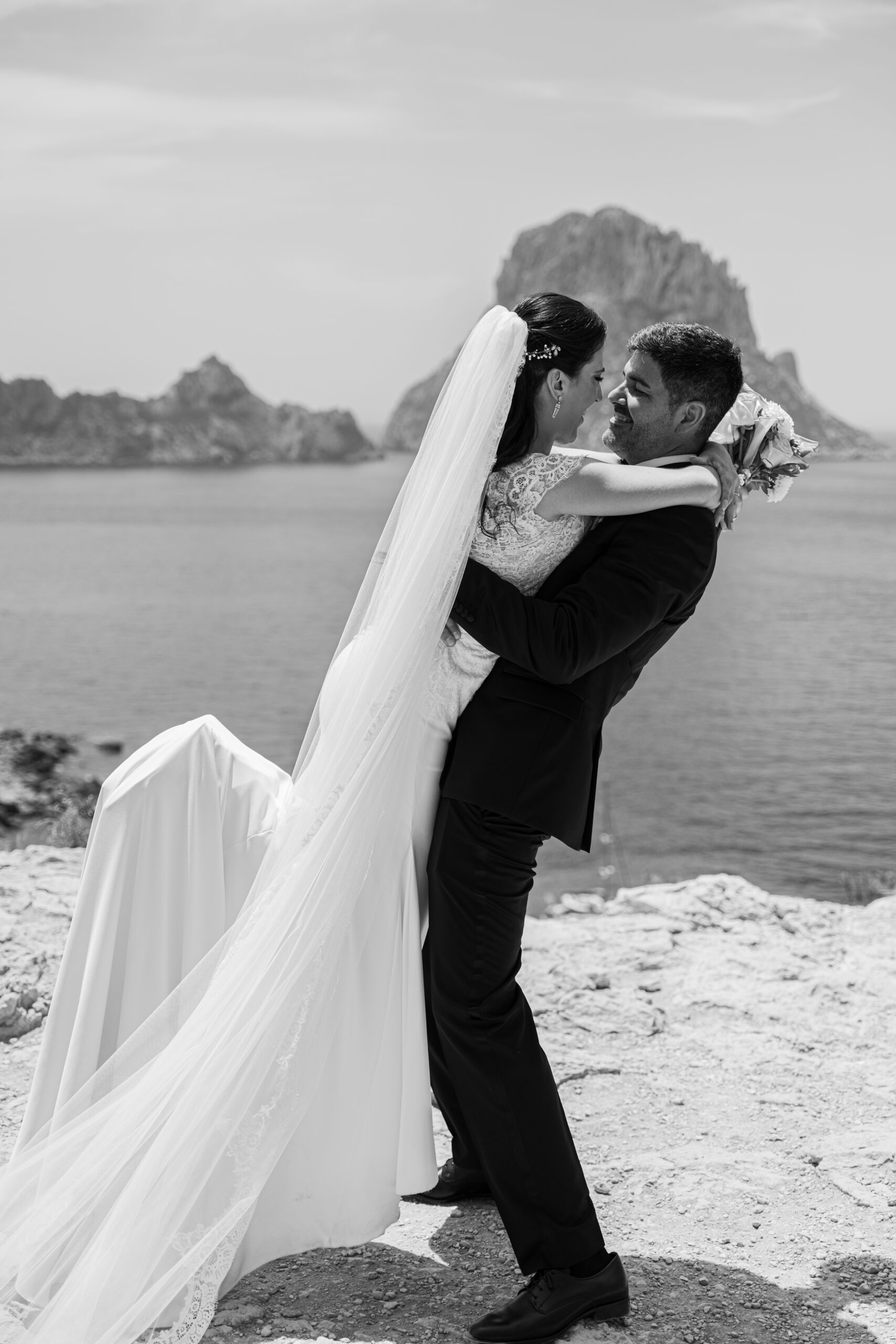 Novios abrazados junto al mar con un peñasco al fondo en una boda al aire libre.