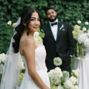 Novia sonriente con vestido blanco y velo, novio con esmoquin negro, fondo de plantas y flores blancas.