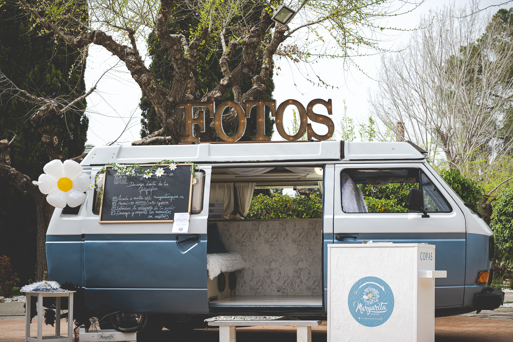 PORTADA Furgoneta vintage azul adaptada como fotomatón con decoración floral y cartel de fotos en un entorno al aire libre.