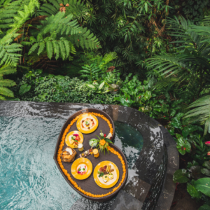 Bandeja con desayuno flotando en piscina rodeada de plantas tropicales.