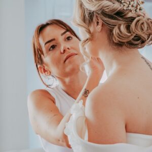 Maquilladora aplicando maquillaje a una mujer de espaldas con peinado recogido y adorno floral.