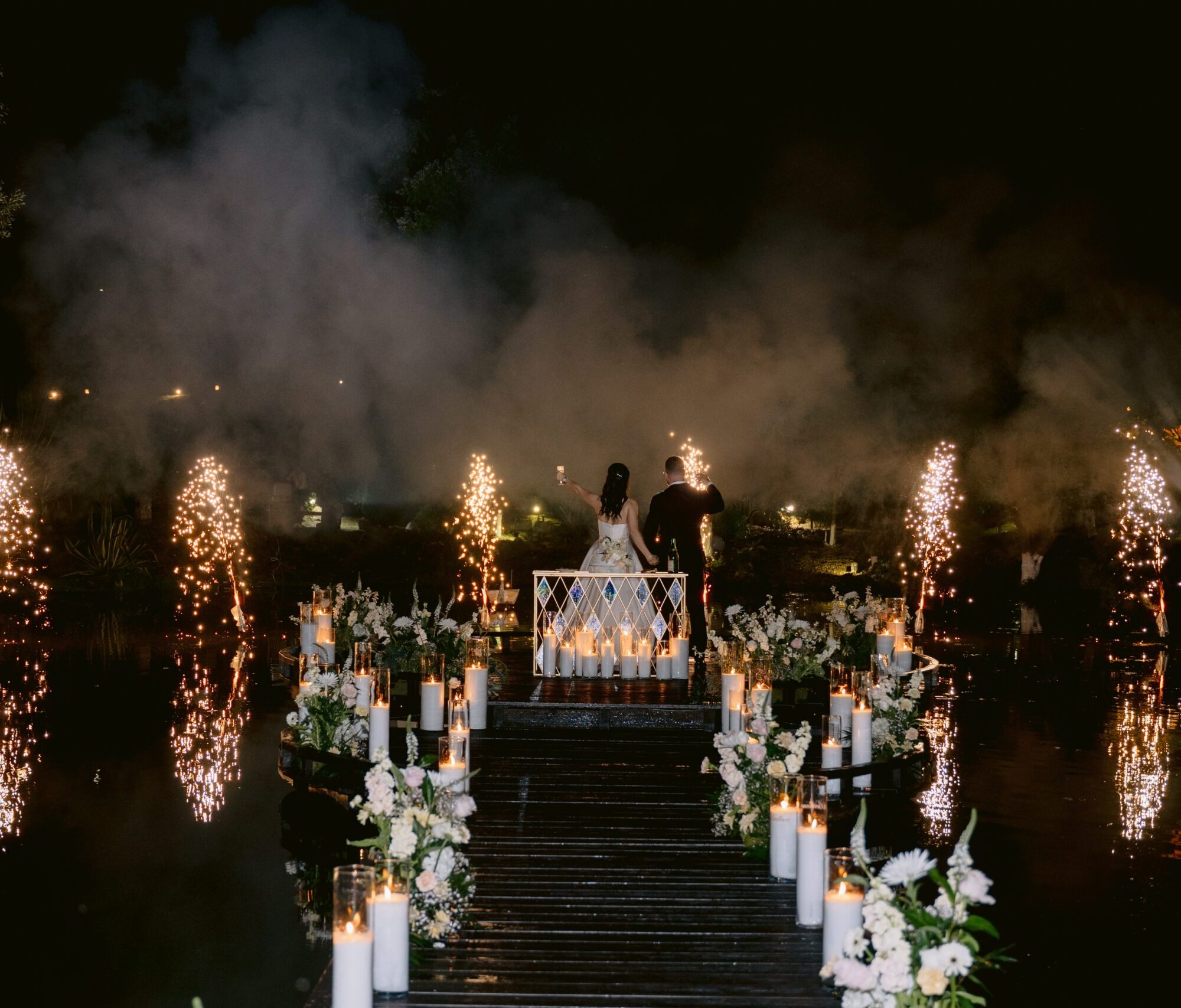 wedding-casal-fotografias-casamento-934 Pareja de novios en muelle decorado con flores y velas, rodeados de fuegos artificiales nocturnos.