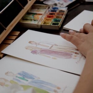 Mano pintando con acuarelas bocetos de vestidos sobre papel blanco.