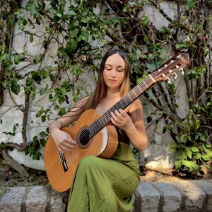 Mujer sentada tocando guitarra clásica frente a un muro cubierto de enredaderas.
