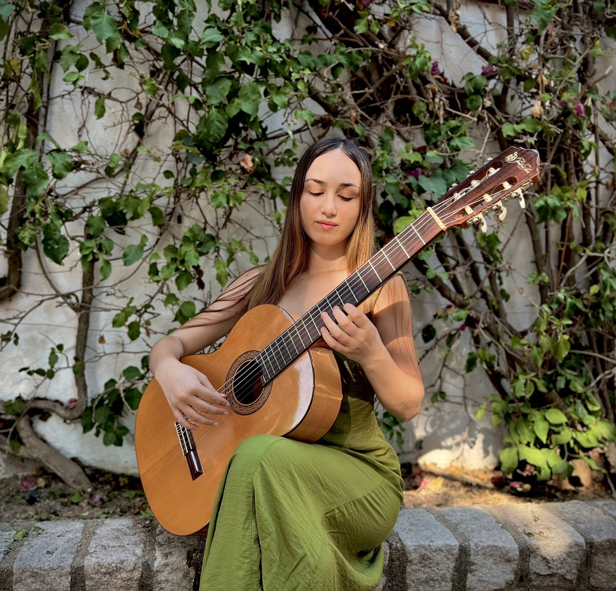Foto Portada Claudia Dorado Mujer sentada tocando guitarra clásica frente a un muro cubierto de enredaderas.