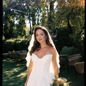 Novia con vestido blanco y ramo de flores en jardín soleado con árboles y sillas al fondo.