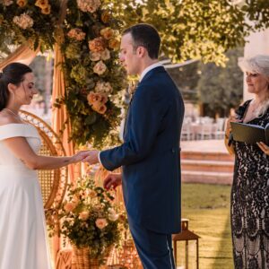 Pareja de novios tomados de la mano frente a una oficiante en ceremonia al aire libre con decoración floral.