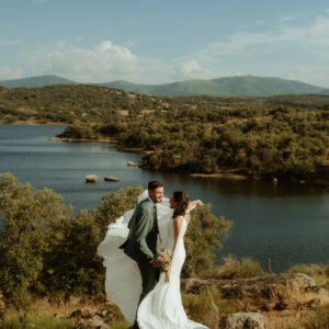 Pareja de novios abrazados junto a un lago rodeado de vegetación y montañas al fondo.