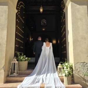 Novios entrando por una puerta grande con vestido de novia largo y capa blanca.
