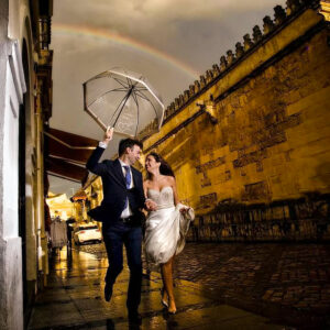 Pareja vestida de boda camina bajo un paraguas con un arcoíris sobre un callejón empedrado al atardecer.