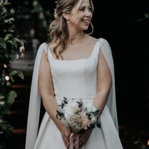 Mujer con vestido blanco de novia y ramo de flores, sonriente, en un entorno oscuro y natural.