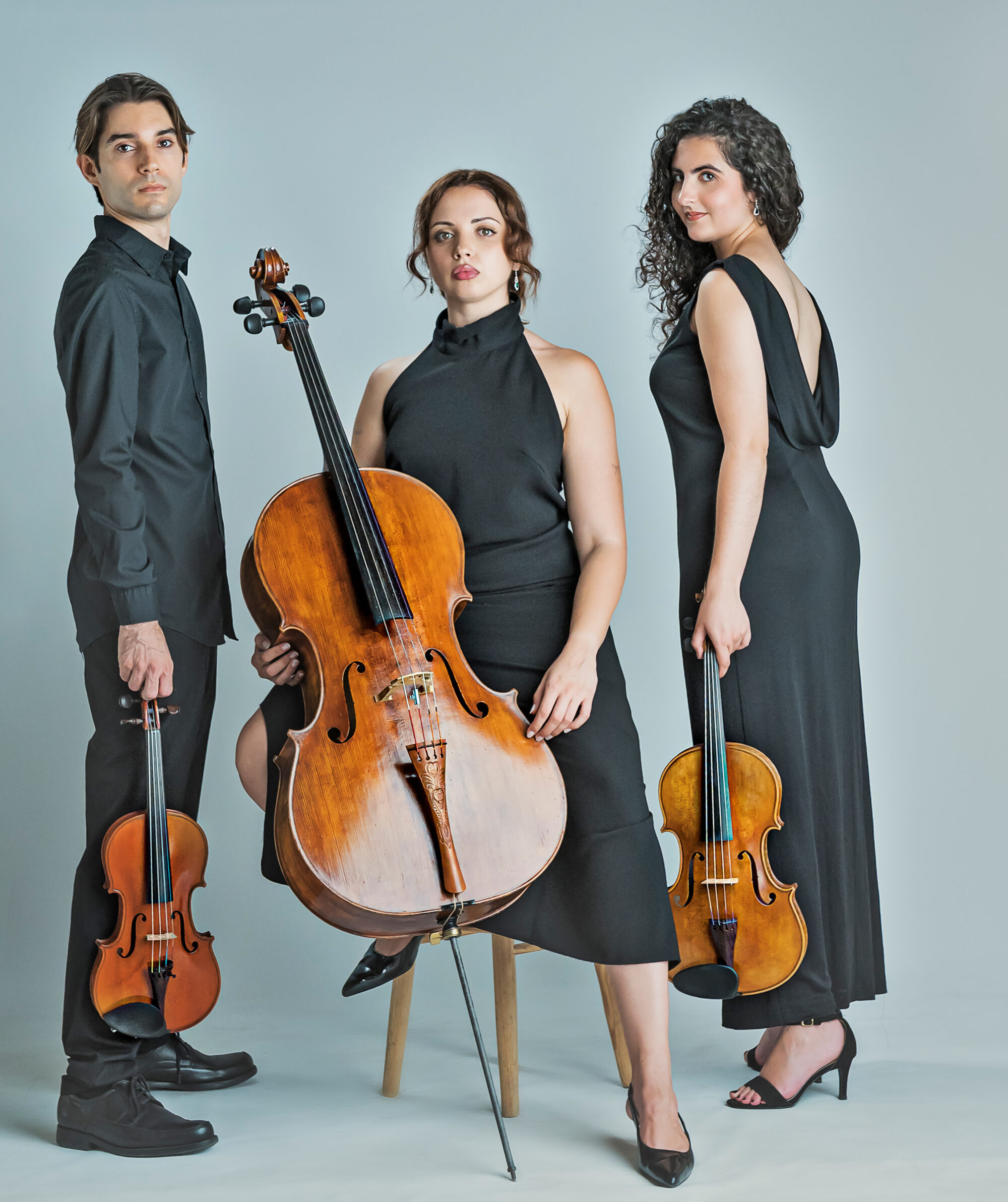Galart fotógrafos Tres músicos con vestidos negros, uno con violonchelo y dos con violines, posan frente a fondo gris.
