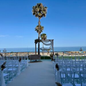 Ceremonia de boda al aire libre con sillas blancas y altar frente al mar y palmeras bajo cielo despejado.