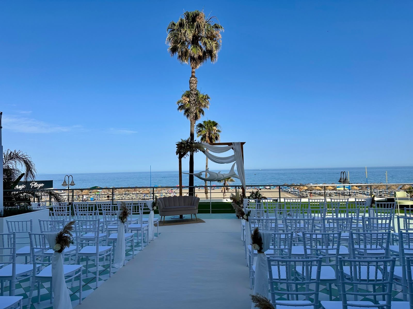0362bf3d-8dc4-4deb-a34d-deb46df5cc52 Ceremonia de boda al aire libre con sillas blancas y altar frente al mar y palmeras bajo cielo despejado.