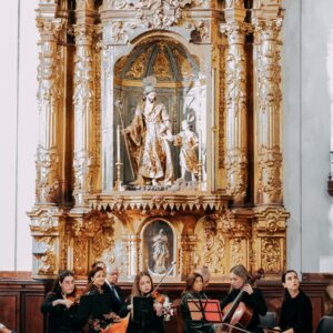 Grupo de músicos con instrumentos de cuerda tocando frente a un retablo barroco tallado en madera.