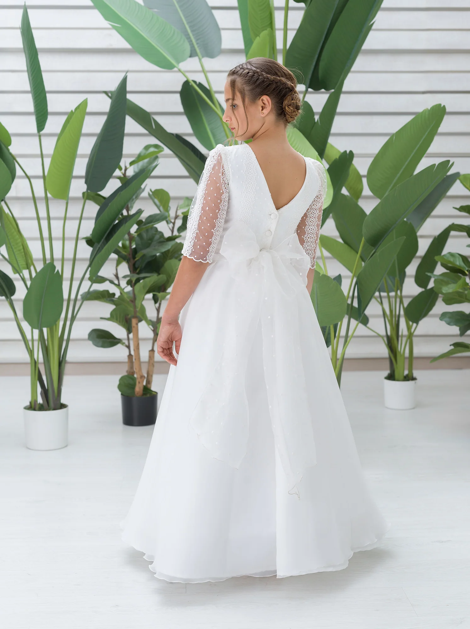 187329 Niña de espaldas con vestido blanco largo y mangas de tul, en fondo con plantas verdes.