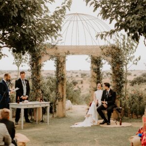 Ceremonia de boda al aire libre con pareja sentada y oficiante bajo estructura con enredaderas.