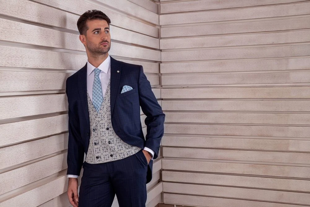 6702 Hombre con traje azul marino, chaleco gris con estampado y corbata celeste apoyado en pared de madera clara.