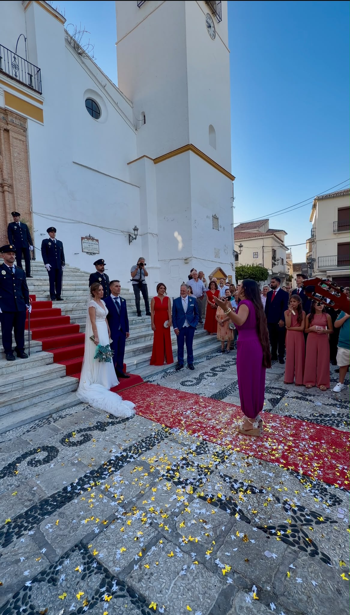 84422776-ecf6-4ed7-a748-6769c3763120 Novios bajando escaleras de iglesia mientras invitados lanzan pétalos y una cantante interpreta una canción.