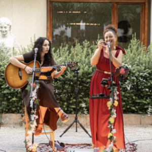 Dos mujeres cantan y tocan guitarra en un jardín decorado con flores y luces.