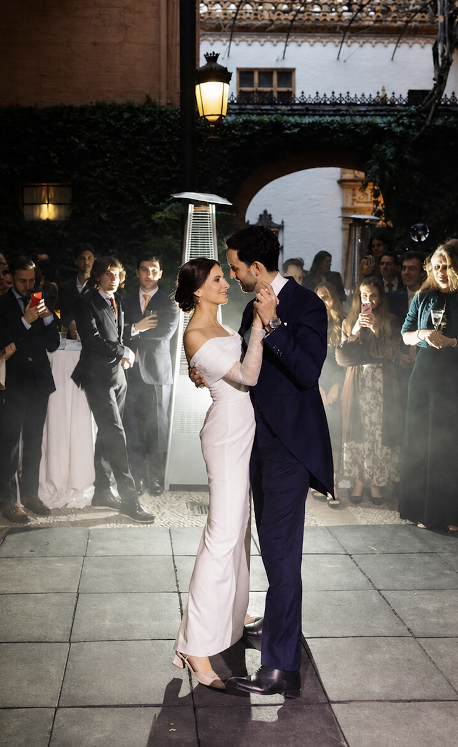 Captura de pantalla 2026-09-03 a las 19.59.05 Pareja bailando en su boda rodeada de invitados en un patio con iluminación cálida.