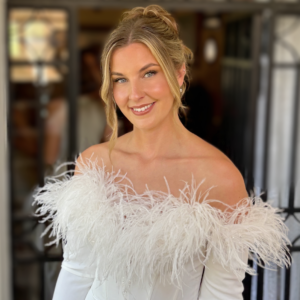 Mujer sonriente con vestido blanco de hombros descubiertos y detalles de plumas, peinado recogido con mechones sueltos.