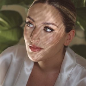 Mujer con maquillaje natural y luz que proyecta sombras de hojas sobre su rostro.