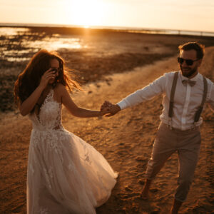 Pareja vestida de boda caminando de la mano al atardecer en la playa.