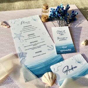 Menú, tarjeta con nombre y nota sobre tela con decoración marina y flores azules secas.