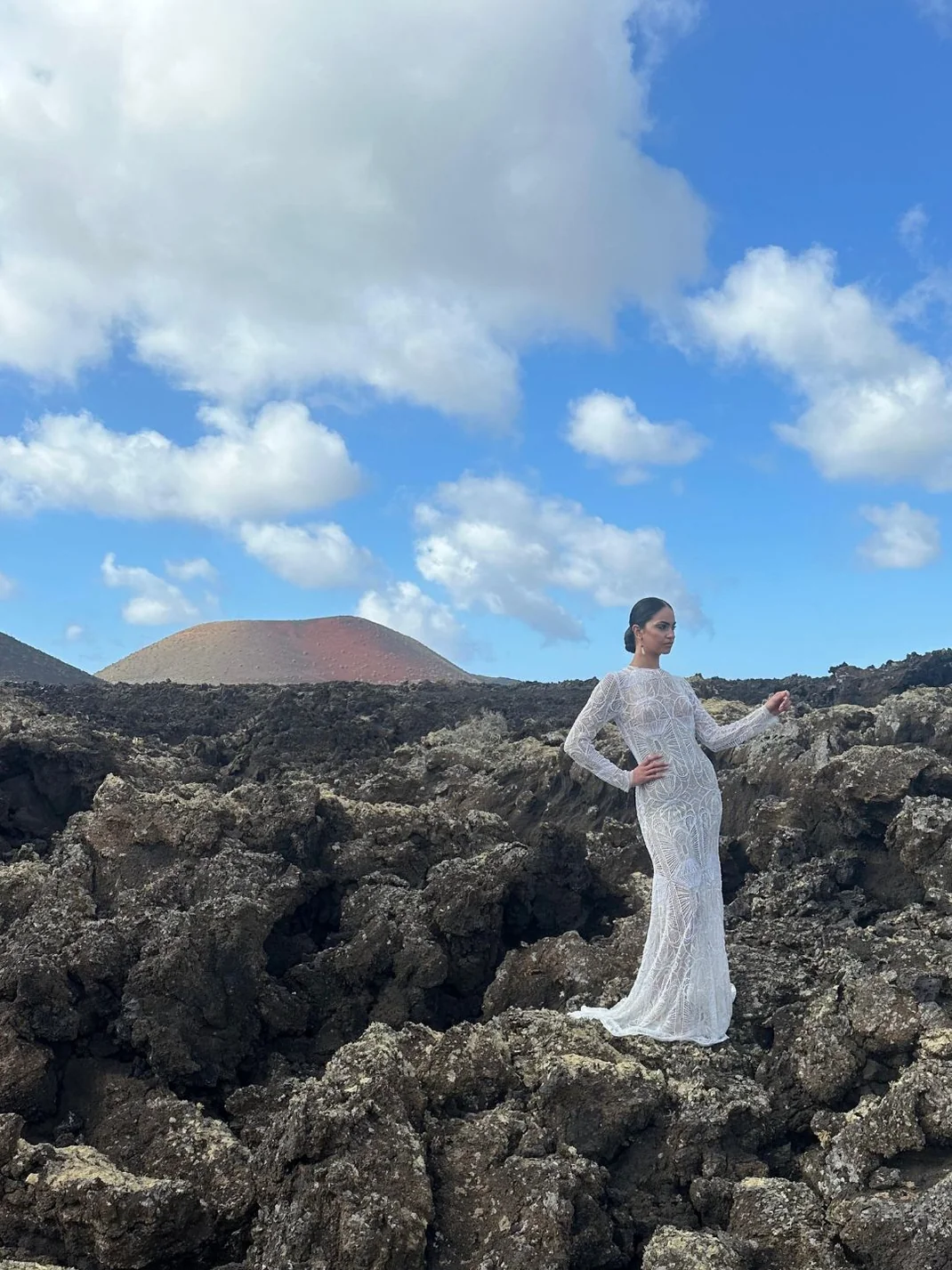 Diseno_sin_titulo_1 Mujer con vestido blanco largo sobre rocas volcánicas bajo cielo azul con nubes.