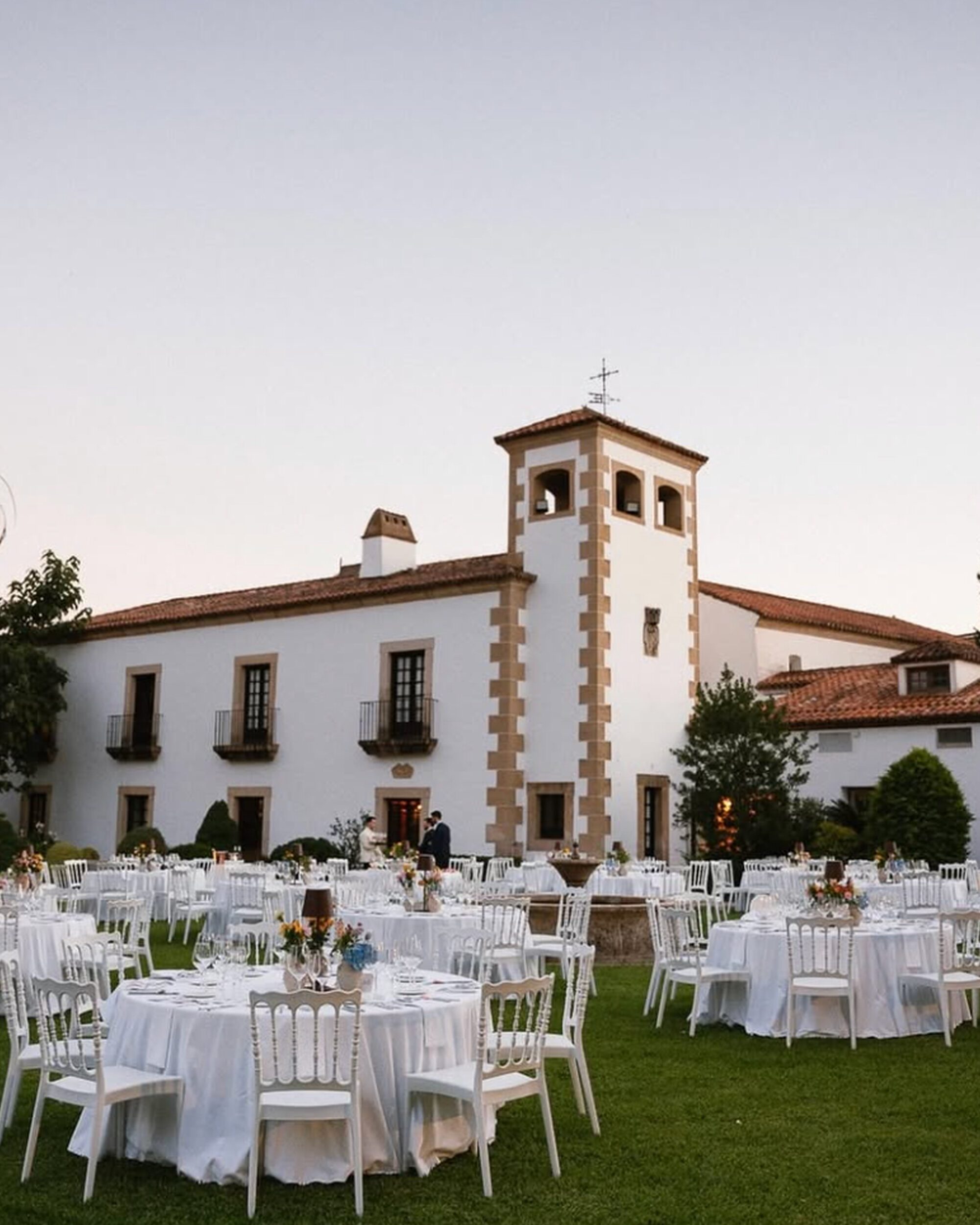 Finca-boda-2500x2500 Jardín con mesas y sillas blancas frente a un edificio histórico de dos plantas con torre central.