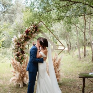 Pareja de novios abrazados bajo un arco floral en un entorno natural con árboles y césped.