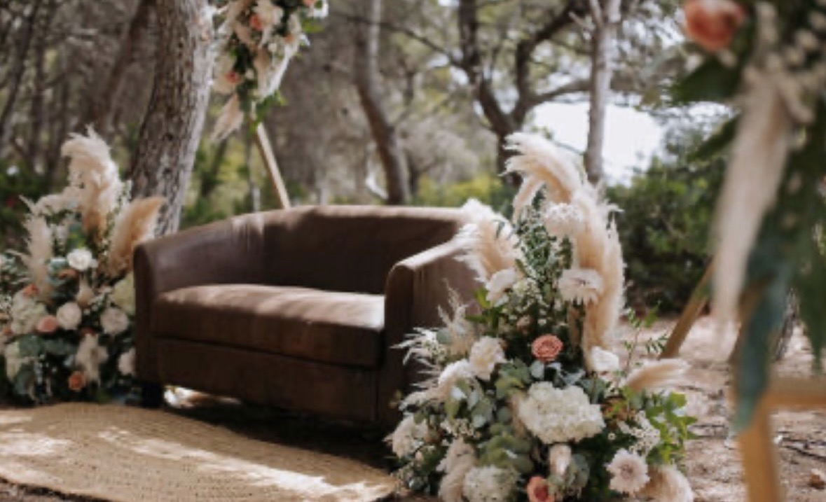 Screenshot Sofá marrón rodeado de arreglos florales en un entorno natural con árboles.