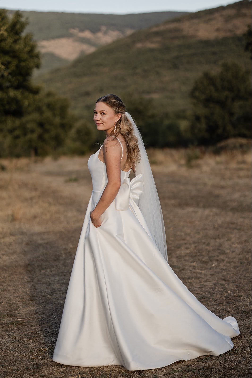 PHOTO-2026-09-08-18-21-54 Mujer de perfil con vestido de novia blanco y velo largo en un paisaje natural con colinas y árboles.