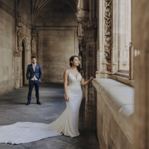 Novia con vestido largo blanco junto a ventana, novio de traje azul al fondo en pasillo antiguo con arcos.