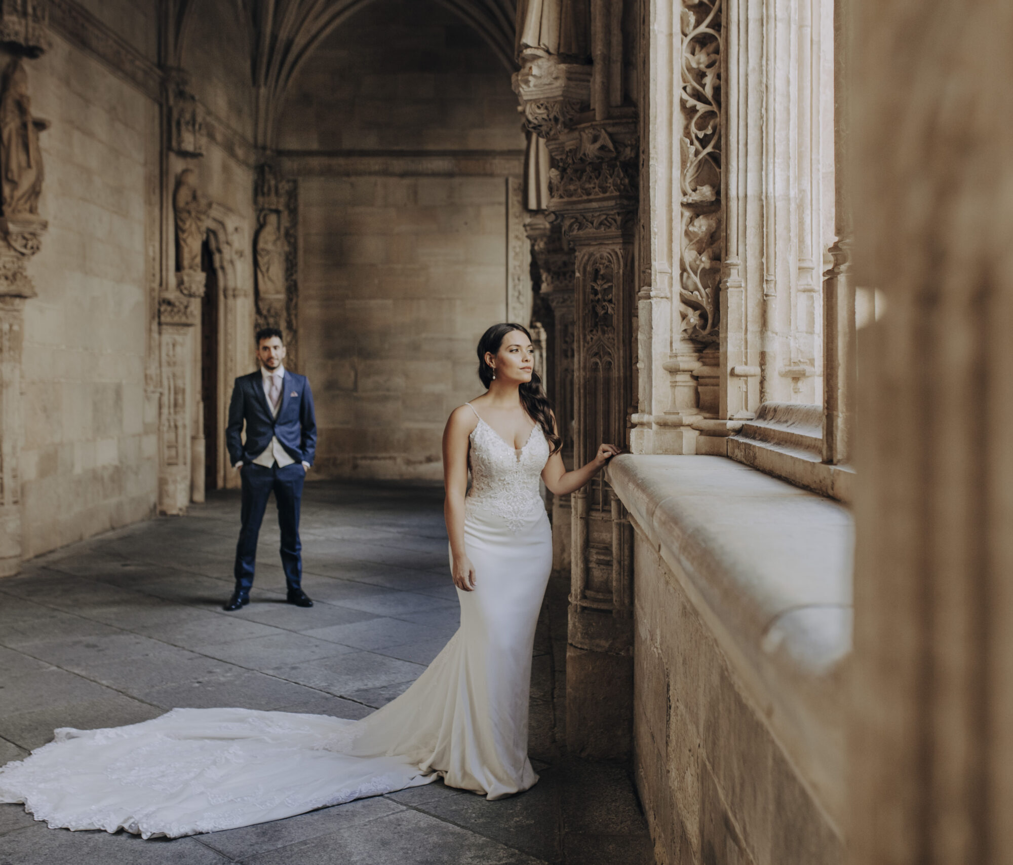 PosbodaSonia&Jaime-31 Novia con vestido largo blanco junto a ventana, novio de traje azul al fondo en pasillo antiguo con arcos.