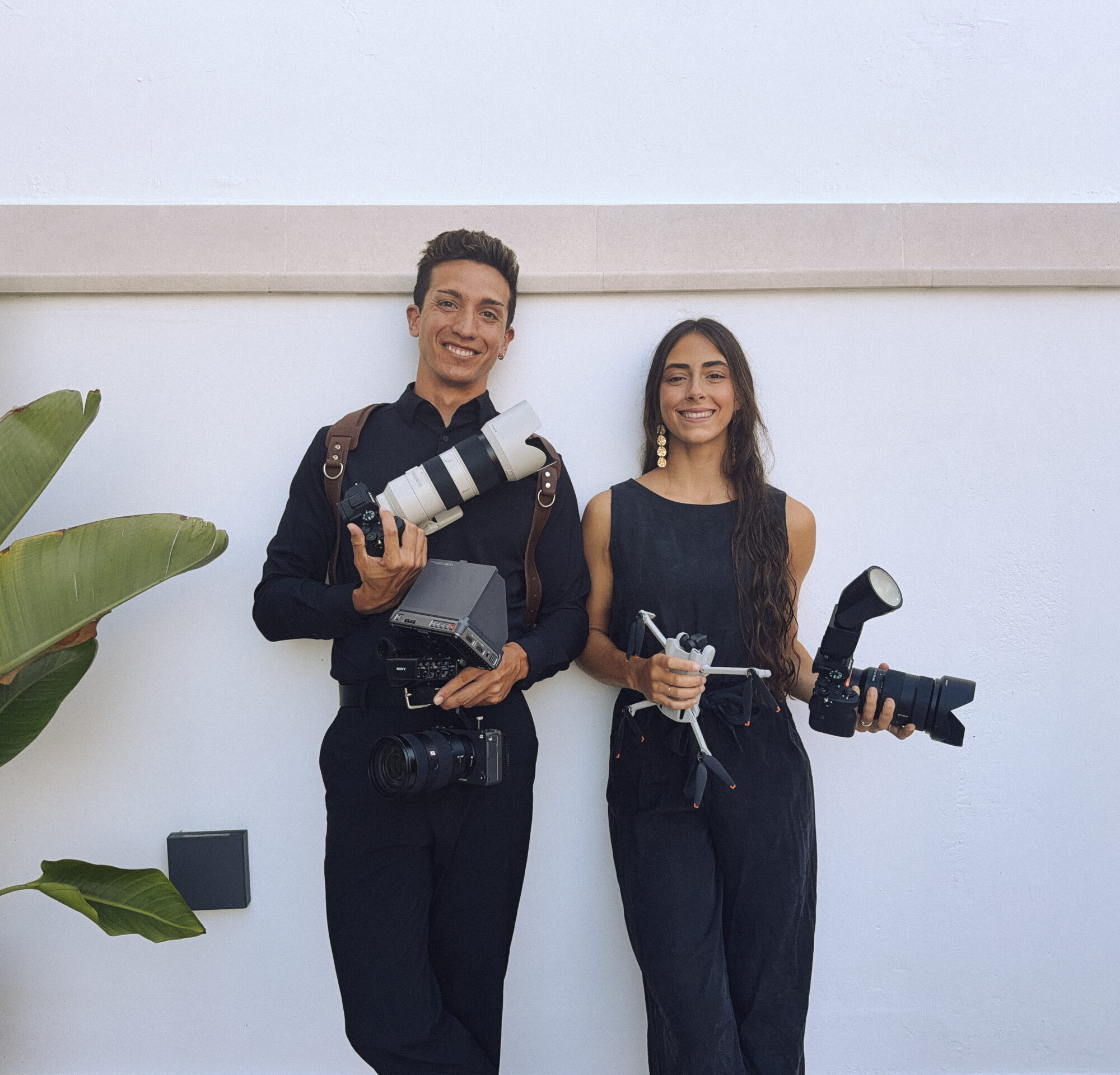 TEAMBRIDE 1 Pareja sonriente con cámaras fotográficas y equipo de fotografía frente a pared blanca.