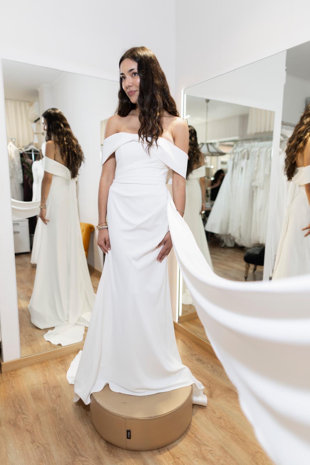c32a5b68-73a3-4d38-b3cd-af92f85978a0 Mujer con vestido blanco largo de hombros descubiertos y abertura lateral, posando frente a un espejo en un atelier.